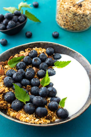 Homemade granola with yogurt, blueberries and mint leaves in a bowl on a turquoise background, delicious healthy breakfastの写真素材