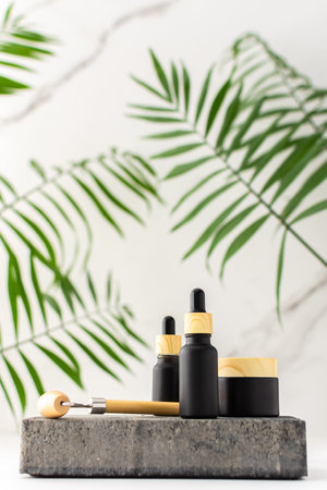 Concrete black podium for cosmetic products close-up among tropical leaves against the background of a marble white wall, bottle with a pipette and a jar of cream and a massage roller for the faceの写真素材