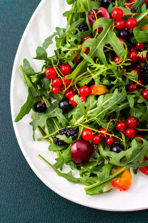 Summer delicious salad with arugula and fresh berries, red and black currants, sweet cherries and black mulberries, salad in an oval white plate on a green backgroundの写真素材