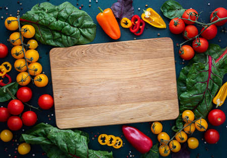 Empty cutting board in the middle of fresh organic vegetables on a green background, food background, farm products, colorful peppers and tomatoes, chard leaves and onionsの写真素材