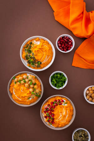 Pumpkin puree soup of butternut squash in bowls with chickpeas and green onions, pumpkin and pomegranate seeds, top view on plates with food on brown backgroundの写真素材