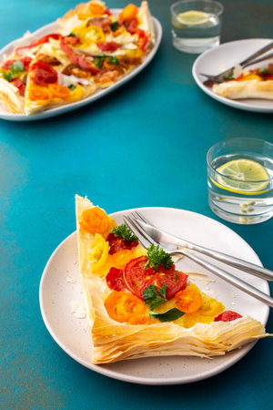 Portion of puff pastry pie with colorful tomatoes, parsley, basil and mozzarella in plates on the table, delicious breakfast, top viewの写真素材