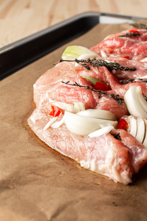 A large piece of raw pork meat prepared for baking, with onions and spices and thyme on parchment paper on a baking sheetの写真素材