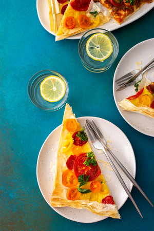 Portion of puff pastry pie with colorful tomatoes, parsley, basil and mozzarella in plates on the table, delicious breakfast, top viewの写真素材