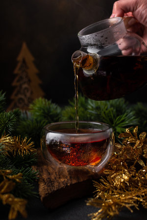 Woman pouring hot brewed black tea in a glass teapot into a glass mug, cozy winter evening with tea and fir branches and gold Christmas decorの写真素材