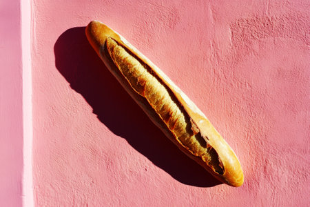 The warm, golden tones of a baguette against a pink wall, with a strong, dramatic shadow adding contrast, bringing out the rustic charm and artisanal quality of the breadの素材