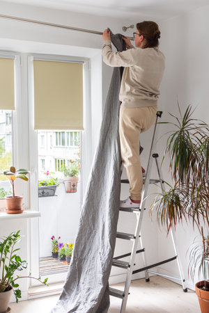 A woman stands on a stepladder and hangs gray curtains on a curtain rod in an apartmentの写真素材