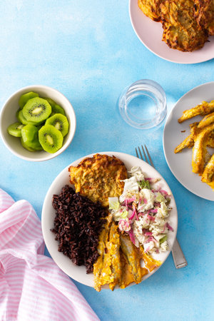 A healthy breakfast featuring brown rice with baked white fish, zucchini-carrot pancakes, cabbage salad with radish, and a refreshing kiwi dessert for a balanced, nutritious start to the dayの写真素材