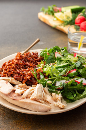 Delicious healthy breakfasts, chicken breast with red rice and salad mix with fresh seasonal organic vegetables and microgreens, a plate with food and a cutting board with vegetablesの写真素材