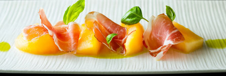 Fresh slices of prosciutto are paired with sweet cantaloupe, garnished with basil leaves, creating a vibrant presentation perfect for summer meals at Italian gatherings.の素材