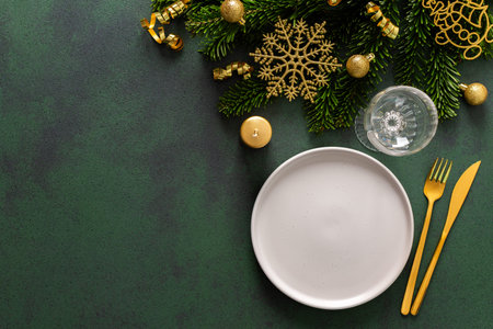 A Christmas table setting with an empty plate and cutlery on a green background. Spruse branches and...
