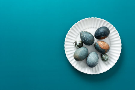 Decorative Easter eggs with various textures and colors are arranged neatly on a white plate, resting on a vibrant teal surface, creating a festive spring atmosphere.の写真素材