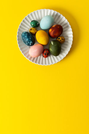 A white plate holds an assortment of vibrant Easter eggs in various colors, placed on a sunny yellow backdrop, perfect for a festive spring gathering.の写真素材