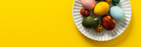 A white plate holds an assortment of vibrant Easter eggs in various colors, placed on a sunny yellow backdrop, perfect for a festive spring gathering, bannerの写真素材