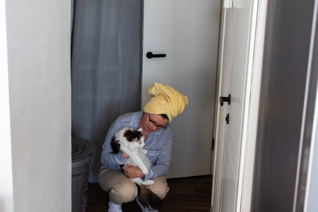 A person is sitting on the floor of a hallway holding a white and brown cat. They have a towel on their head and are dressed in casual clothes. It is daytime and bright inside.の写真素材