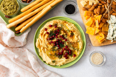 Delight in a classic hummus dish topped with pomegranate seeds, served alongside a variety of cheeses, nuts, and breadsticks perfect for sharing during gatherings.の写真素材