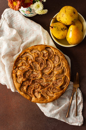A fresh pear pie is placed on a cloth on a table. Nearby, a bowl holds ripe pears and a small vase has flowers. The setting looks warm and inviting for sharing a treat.の写真素材