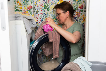 Woman stands in bathroom putting clothes into washing machine while surrounded by towels and laundry, carrying out daily household task with focus and careの写真素材