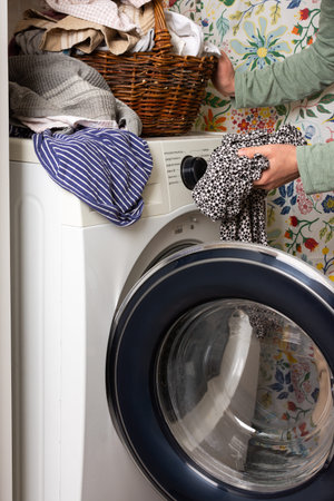A woman sorts and loads laundry into a washing machine. The vibrant floral wall adds warmth to the cozy room. A basket of clothes sits nearby, ready to be washedの写真素材