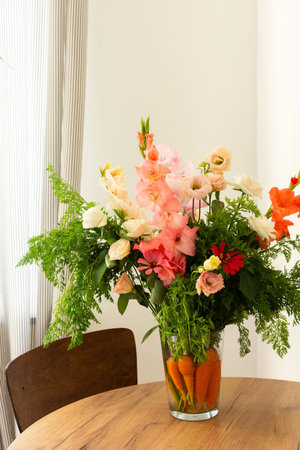A bouquet features roses, gladioli, zinnias, and eustomas. Carrots with green tops sit in water, placed on a wooden table in a bright roomの写真素材