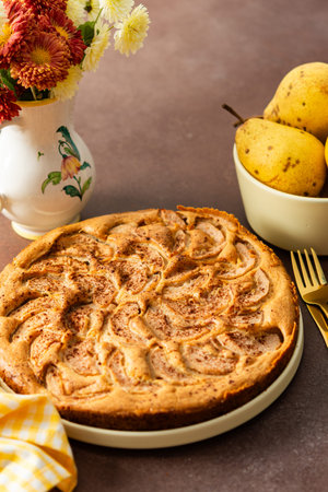 A fresh pear pie is placed on a cloth on a table. Nearby, a bowl holds ripe pears and a small vase has flowers. The setting looks warm and inviting for sharing a treatの写真素材