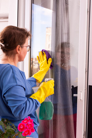 A 50-year-old woman in yellow gloves cleans windows on her balcony, ensuring a clear view before...