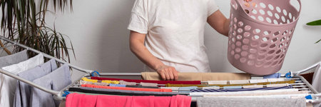 A 50-year-old woman hangs freshly washed laundry on a drying rack inside her room. She is holding a...