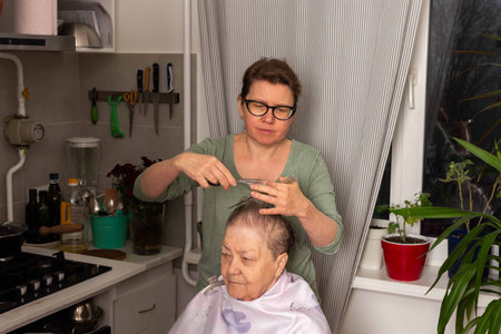 A woman is cutting her mother hair in the kitchen while they enjoy time together. The setting features plants and flowers, creating a lively atmosphere for this home activityの写真素材