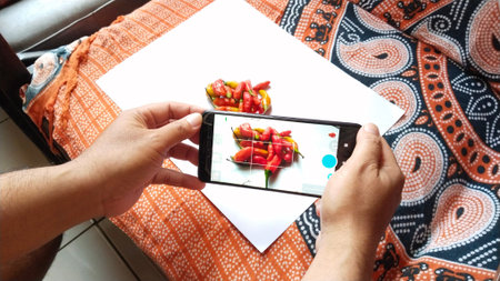 Hands hold a smartphone capturing a vibrant flat lay of red and yellow chili peppers on a white...