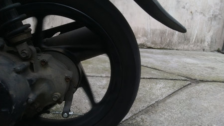 Dirty black motor scooter wheel and engine parts parked on concrete pavement with a weathered wall...