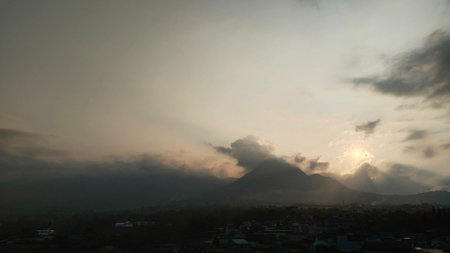 A hazy sunset casts dramatic sunbeams through clouds over a silhouetted mountain range and a town...