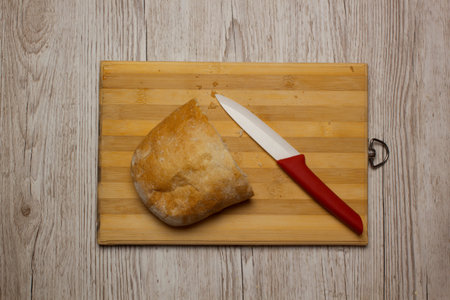 A half of ciabatta bread on a wooden kitchen desk with ceramic knife with red handle