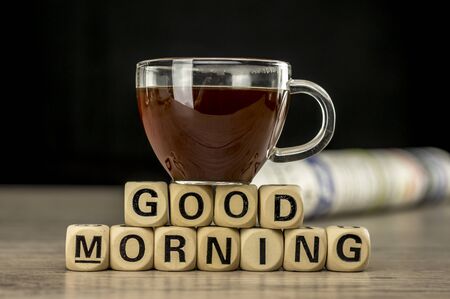 Close up shot of a glass cup of coffee and newspaper. Good morning phrase assembled with wooden letter blocksの写真素材