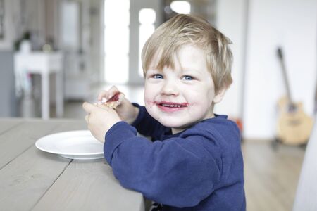a happy blond boy sitting at a table and eating a jam sandwichの写真素材