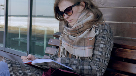 young, beautiful, lovely woman sitting on bench in park, in hands of her notebook. She is wearing a coat. She makes notes and looks very romantic.の写真素材
