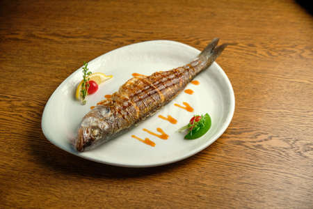 Fried fish with lemon and herbs on a white plate on a wooden tableの写真素材