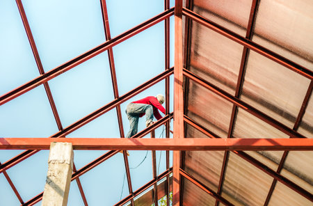 Roofer working in special protective work wear gloves, using air or pneumatic nail gun installing asphalt shingle on top of the new roof, Concept of residential building under construction.の写真素材