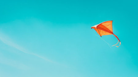 A red kite flying high in the sky, with its tail fluttering in the wind against a bright blue background.の素材