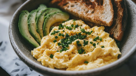 A comforting bowl of creamy scrambled eggs topped with chives, served with crispy toast and sliced avocado on a bright breakfast table.の素材