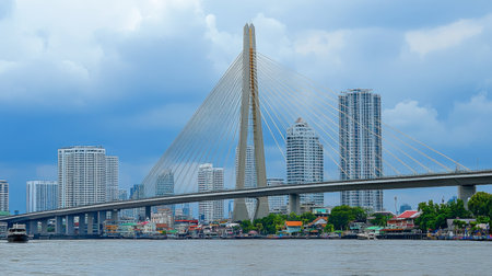 A striking modern bridge stands tall against an urban skyline, reflecting off the river water. Dramatic clouds enhance the beauty of this vibrant cityscape.の素材