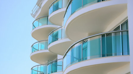 An elegant view of a modern building showcasing curved balconies with glass railings against a clear blue sky, highlighting contemporary architectural design elements.の素材