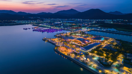 Captivating aerial view of a seaside city during dusk, showcasing beautiful illuminated buildings, boats in the harbor, and serene mountains in the background.の素材