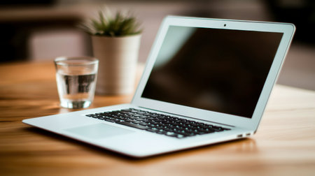 A stylish laptop rests on a wooden table beside a glass of water and a small potted plant, creating an inviting and modern workspace ideal for productivity and creativity.の素材