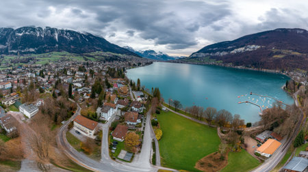 Stunning aerial perspective showcasing a serene lake surrounded by mountains and a picturesque village under an overcast sky, ideal for nature enthusiasts and travel planners.の素材