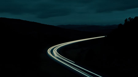 A captivating long exposure image showcasing car lights illuminating a winding road at night. The serene landscape features dark mountains and a cloudy sky, creating a moody ambiance.の素材