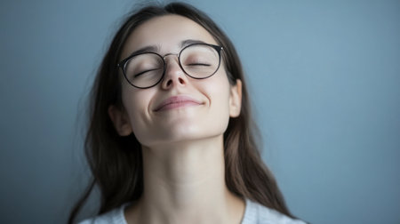 A serene young woman with glasses smiles softly, exuding joy and calm against a simple blue background. Ideal for themes of happiness and confidence.の素材