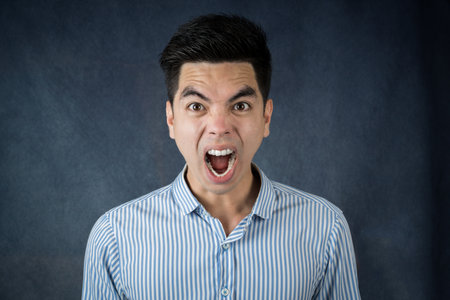 Portrait stressed and  screaming crazy handsome young asian man wearing a blue shirt isolated on dark wall background.  Asia people.の写真素材