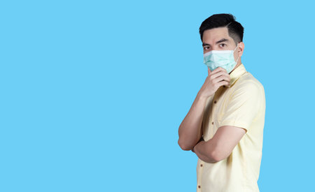 Stressful COVID-19 Coronavirus portrait handsome young asian man wearing yellow shirt and mask protection from covid 19 isolated on blue background in studio. Asian man people. COVID-19 concept.の写真素材