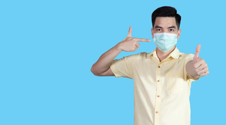 COVID-19 Coronavirus portrait handsome young asian man wearing yellow shirt and mask protection from covid 19 and thumbs up sign isolated on blue background in studio. Asian people. COVID-19 concept.の写真素材