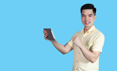 Portrait handsome young asian man wearing a yellow shirt holding smart phone or tablet feeling celebrating or successful isolated on blue background. Asia people.の写真素材